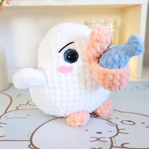 May include: White crocheted plush toy with a pink beak and blue fish in its mouth. The toy has large black eyes and pink cheeks.