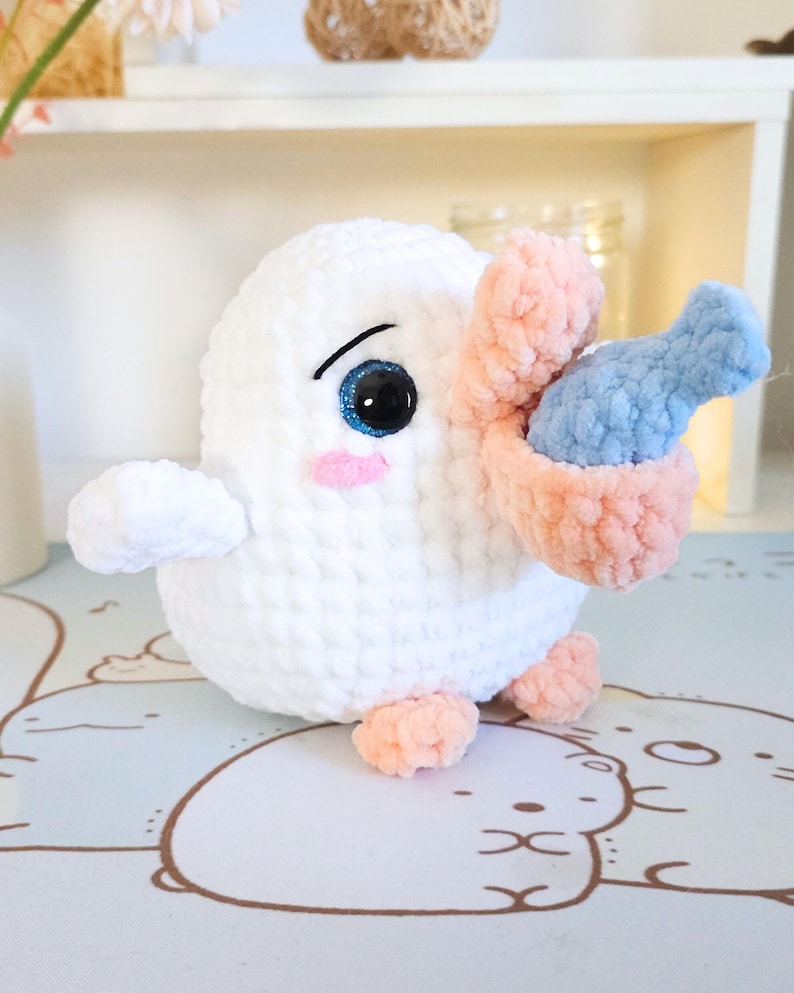 May include: White crocheted plush toy with a pink beak and blue fish in its mouth. The toy has large black eyes and pink cheeks.