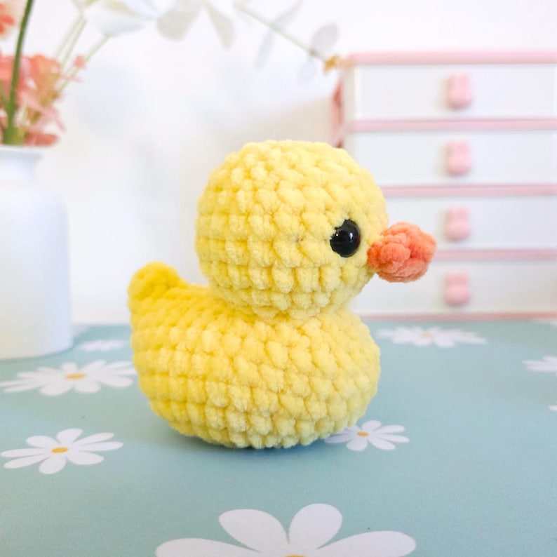 Puede incluir: Un pato amarillo tejido a ganchillo, con una textura suave. El pato tiene un peque&ntilde;o pico naranja y ojos negros. El pato est&aacute; sobre una superficie azul claro con patrones de margaritas blancas. Un jarr&oacute;n blanco con flores rosas en el fondo.
