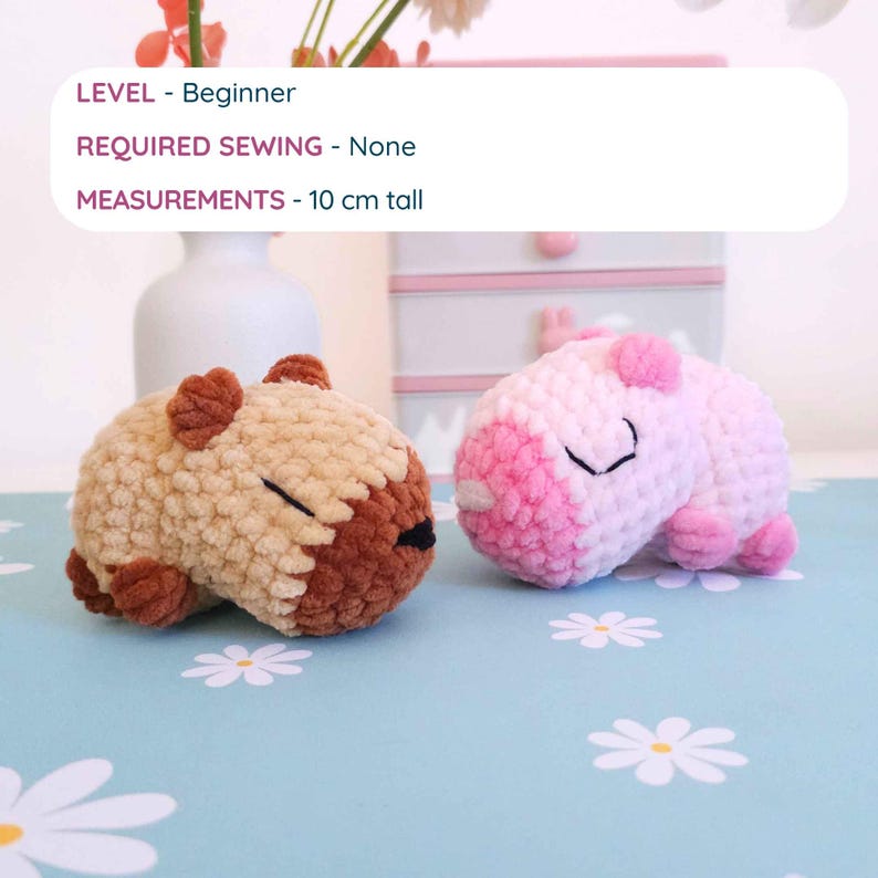 Pu&ograve; includere: Due peluche di capibara all'uncinetto, uno beige e marrone, l'altro rosa e bianco. Il capibara beige ha la faccia e le orecchie marroni, mentre quello rosa ha dettagli rosa. Il testo sull'immagine dice "LIVELLO - Principiante", "CUCITO RICHIESTO - Nessuno", e "MISURE - 10 cm di altezza".