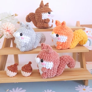 Op de afbeelding: Een verzameling handgemaakte gehaakte eekhoorns in verschillende kleuren, waaronder bruin, grijs, oranje en roze, met witte en roze accenten. Er zijn ook twee kleine gehaakte eikels aanwezig. De eekhoorns worden tentoongesteld op een houten plank.