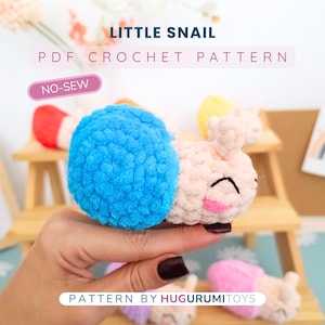 May include: A crocheted snail toy with a blue shell and a light pink body is held in a hand. The text "LITTLE SNAIL PDF CROCHET PATTERN" is above the snail. The text "NO-SEW" is in a pink oval. The text "PATTERN BY HUGURUMITOYS" is below.