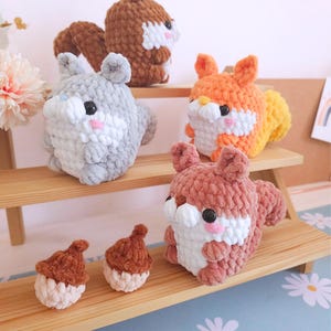 Op de afbeelding: Een verzameling handgemaakte gehaakte eekhoornknuffels in verschillende kleuren, waaronder grijs, oranje en bruin. De eekhoorns hebben witte accenten en zwarte ogen. Er zijn ook twee kleine gehaakte eikels aanwezig. De knuffels worden tentoongesteld op een houten plank.