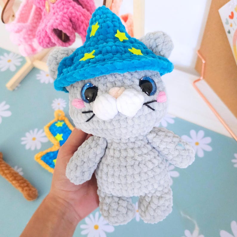 May include: A handmade gray crochet cat wearing a blue wizard hat with yellow stars. The cat has blue glittery eyes, pink cheeks, and a white nose. The cat is holding a blue and yellow butterfly.
