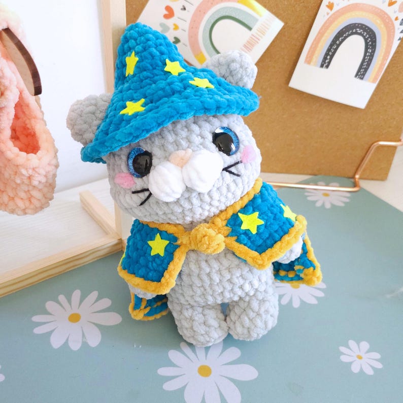 May include: A handmade gray crochet cat wearing a blue wizard hat and cape, both adorned with yellow stars. The cat has black eyes, pink cheeks, and white features. The cape is trimmed with yellow. The cat is sitting on a daisy-patterned surface.