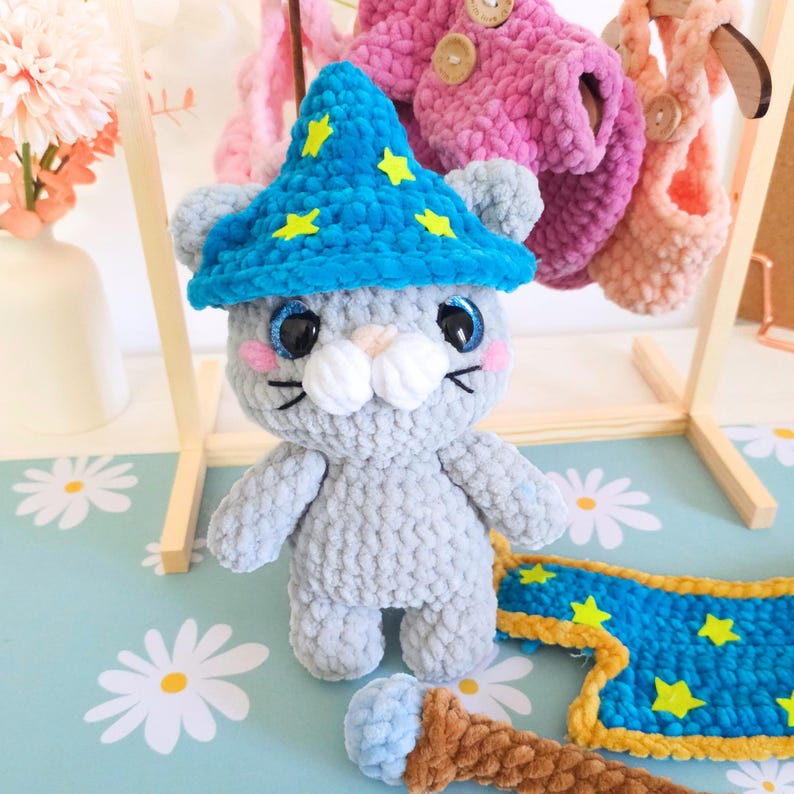 May include: A handmade gray crochet cat wearing a blue wizard hat with yellow stars. The cat has blue eyes, pink cheeks, and white whiskers. A blue and gold magic carpet and a brown and blue wand are also visible.