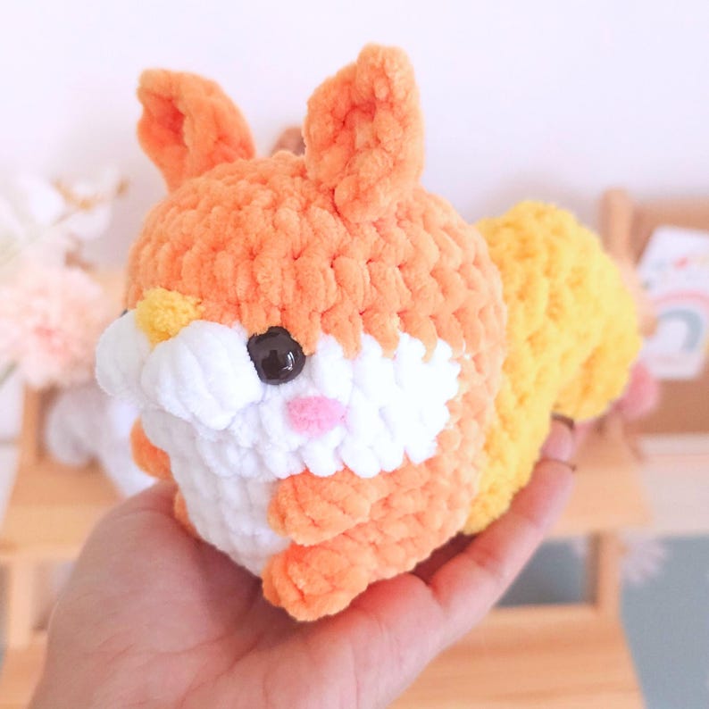 Op de afbeelding: Een handgemaakte, gehaakte eekhoornknuffel in oranje, wit en geel. De eekhoorn heeft een pluizige staart, kleine oren en een witte buik. De knuffel wordt in een hand gehouden, wat de grootte en textuur benadrukt.