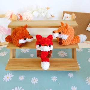 May include: Three handmade crochet fox toys in orange and red with white and brown accents. The foxes have small black eyes and white muzzles. They are displayed on a wooden shelf with a light blue floral background.