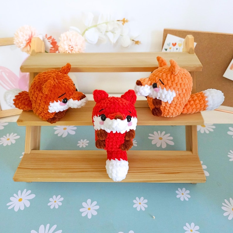 May include: Three handmade crochet fox toys in orange and red with white and brown accents. The foxes have small black eyes and white muzzles. They are displayed on a wooden shelf with a light blue floral background.