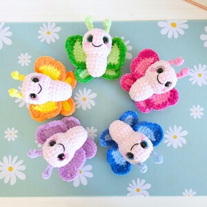 Puede incluir: Cinco mariposas de ganchillo coloridas en rosa, verde, naranja, morado y azul. Cada mariposa tiene un cuerpo blanco, ojos negros y una cara sonriente. Las alas son de varios tonos del mismo color. Las mariposas est&aacute;n dispuestas sobre una superficie azul claro con patrones de margaritas blancas.