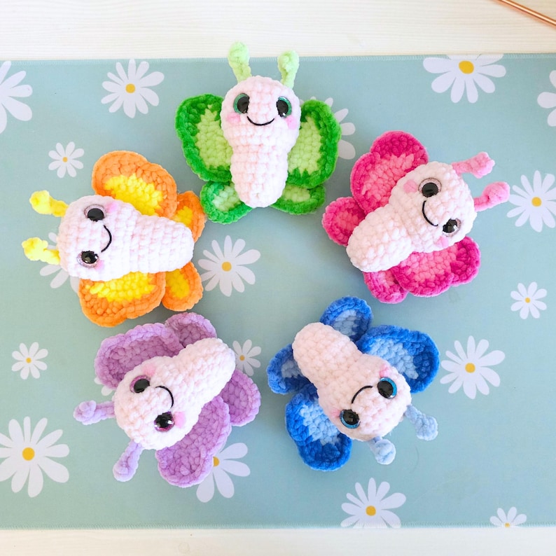 Puede incluir: Cinco mariposas de ganchillo coloridas en rosa, verde, naranja, morado y azul. Cada mariposa tiene un cuerpo blanco, ojos negros y una cara sonriente. Las alas son de varios tonos del mismo color. Las mariposas est&aacute;n dispuestas sobre una superficie azul claro con patrones de margaritas blancas.