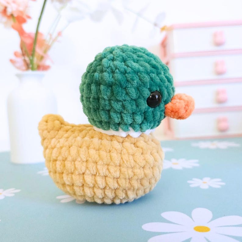 Puede incluir: Un juguete de pato hecho a mano con ganchillo, con cabeza verde, cuerpo amarillo y pico naranja. El pato tiene ojos negros y un cuello blanco. El peluche est&aacute; sobre una superficie azul con motivos de flores blancas.