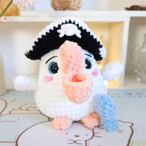 May include: A white crocheted plush toy bird wearing a black and white pirate hat with a skull and crossbones design. The bird has a pink beak and blue eyes. The bird is holding a blue crocheted fish in its right wing.