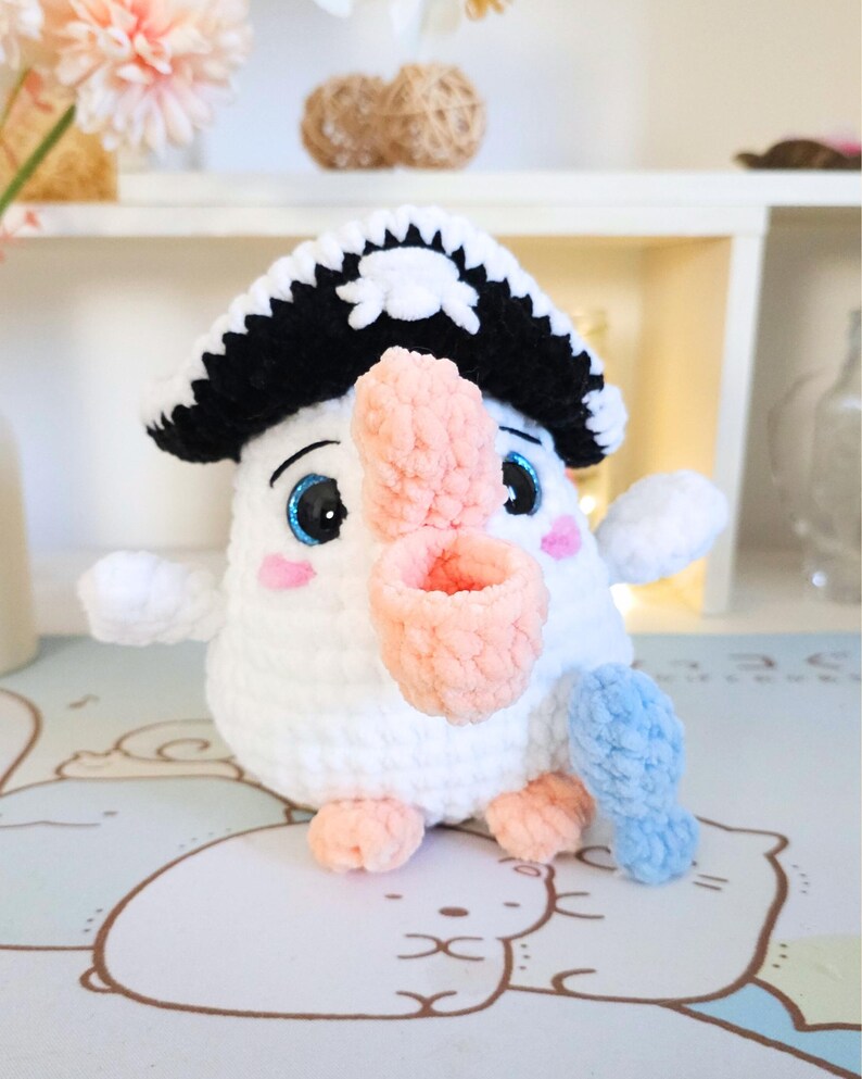 May include: A white crocheted plush toy bird wearing a black and white pirate hat with a skull and crossbones design. The bird has a pink beak and blue eyes. The bird is holding a blue crocheted fish in its right wing.