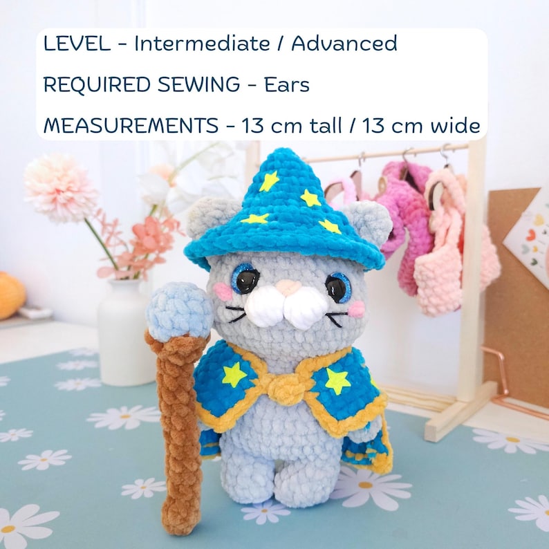May include: A handmade gray cat plushie wearing a blue wizard hat and cape with yellow star accents. The plushie is holding a brown and blue staff. The text on the image reads: LEVEL - Intermediate / Advanced, REQUIRED SEWING - Ears, MEASUREMENTS - 5.1 inches tall / 5.1 inches wide.