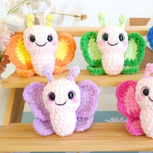 Puede incluir: Una colecci&oacute;n de mariposas de ganchillo hechas a mano y coloridas. Cada mariposa presenta una cara sonriente, ojos grandes y alas en tonos naranja, verde, morado y rosa. Las mariposas se exhiben en una estanter&iacute;a de madera, mostrando su apariencia suave y texturizada.
