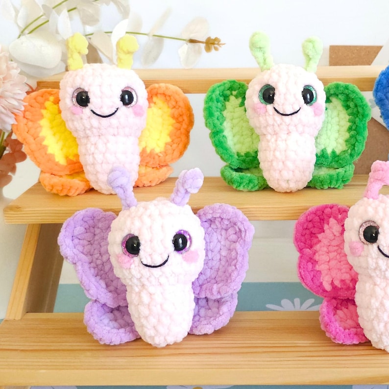 Puede incluir: Una colecci&oacute;n de mariposas de ganchillo hechas a mano y coloridas. Cada mariposa presenta una cara sonriente, ojos grandes y alas en tonos naranja, verde, morado y rosa. Las mariposas se exhiben en una estanter&iacute;a de madera, mostrando su apariencia suave y texturizada.