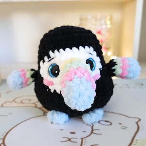 May include: A crocheted toucan plush toy with a black body, white and rainbow coloured beak, and blue eyes. The toucan is sitting on a white surface with a cartoon pattern.