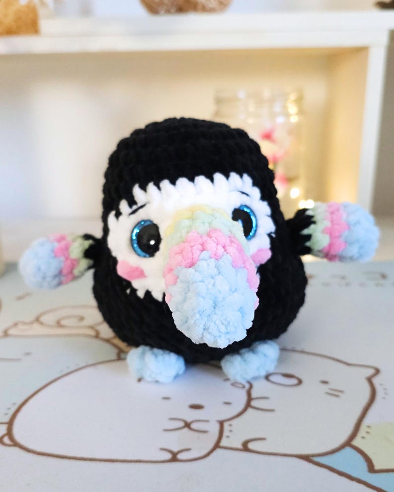 May include: A crocheted toucan plush toy with a black body, white and rainbow coloured beak, and blue eyes. The toucan is sitting on a white surface with a cartoon pattern.