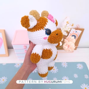 May include: A handmade crochet cow plushie with white and brown spots, a pink bow, and large blue eyes. The plushie has a soft, textured appearance. The text "PATTERN BY HUGURUMITOYS" is visible at the bottom of the image.