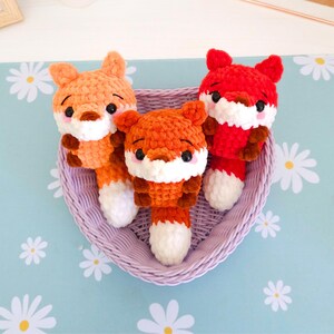 May include: Three crocheted fox toys in a purple woven basket. The foxes are orange and red with white and brown accents. They have black eyes and are arranged on a light blue surface with white daisy patterns.