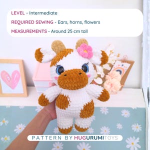 May include: A handmade crochet cow plushie, primarily white with brown spots and accents. It has large blue eyes, a brown muzzle, and a pink flower on its head. The text on the image says "LEVEL - Intermediate" and "PATTERN BY HUGURUMITOYS". The cow is approximately 9.8 inches tall.