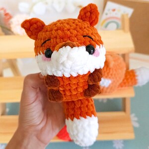 May include: A handmade crochet fox toy in shades of orange, white, and brown. The fox has black eyes, pink cheeks, and a fluffy white chest. The tail is orange with a white tip. The toy is soft and cuddly.