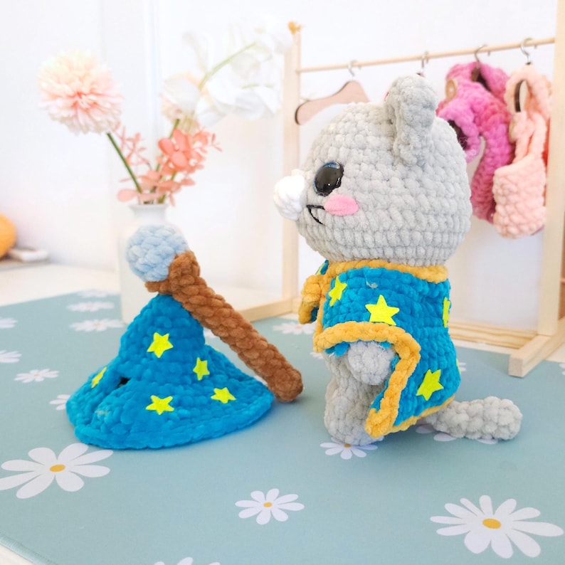 May include: A handmade crochet wizard cat doll with a blue and yellow star-patterned robe and a matching hat. The cat has gray fur, pink cheeks, and a white nose. A brown and blue wand is next to the cat.