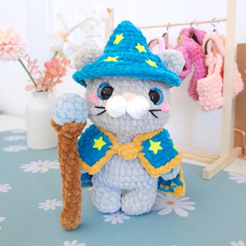 May include: A handmade gray plush toy of a wizard mouse, wearing a blue hat and cape with yellow stars. The mouse holds a brown staff with a blue orb. The toy has black whiskers and pink cheeks. The background includes a floral arrangement and a clothing rack.