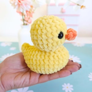 Puede incluir: Un patito amarillo tejido a ganchillo, con cuerpo redondo, ojos negros y pico naranja. El peluche se sostiene en una mano, resaltando su textura suave. El fondo incluye un jarr&oacute;n con flores y una superficie azul claro.
