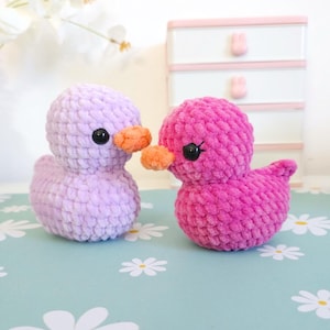 Puede incluir: Dos patitos de ganchillo hechos a mano. Uno es lavanda, el otro rosa intenso. Ambos tienen picos naranjas y ojos negros. Los patitos est&aacute;n sobre una superficie azul claro con motivos de margaritas blancas. El fondo incluye un armario blanco y rosa.