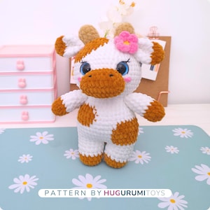 May include: A handmade crochet cow plushie with white and brown spots, a brown muzzle, and a pink flower on its head. The cow has large blue eyes and is standing on a light blue surface with daisy patterns. Text on the bottom reads "PATTERN BY HUGURUMITOYS."