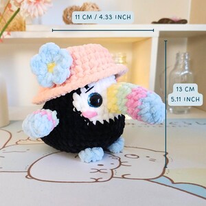 May include: A crocheted toucan plush toy wearing a pink and white crocheted sun hat with a blue flower. The toucan is 13 cm tall and 11 cm wide.