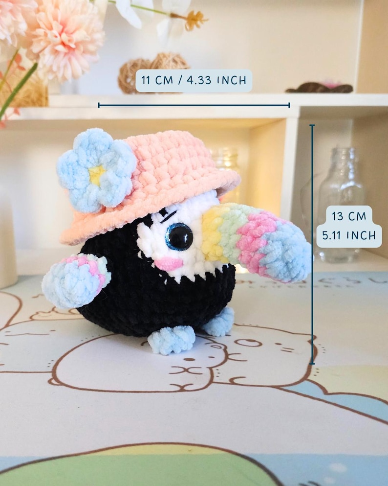 May include: A crocheted toucan plush toy wearing a pink and white crocheted sun hat with a blue flower. The toucan is 13 cm tall and 11 cm wide.