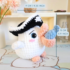 May include: White crocheted amigurumi toy with a black and white pirate hat, holding a pink and blue crocheted object. The toy is 13 cm tall and 11 cm wide.