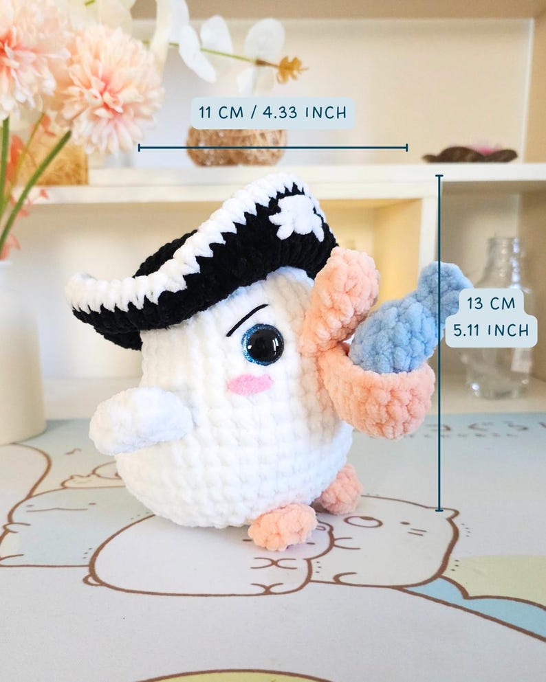 May include: White crocheted amigurumi toy with a black and white pirate hat, holding a pink and blue crocheted object. The toy is 13 cm tall and 11 cm wide.