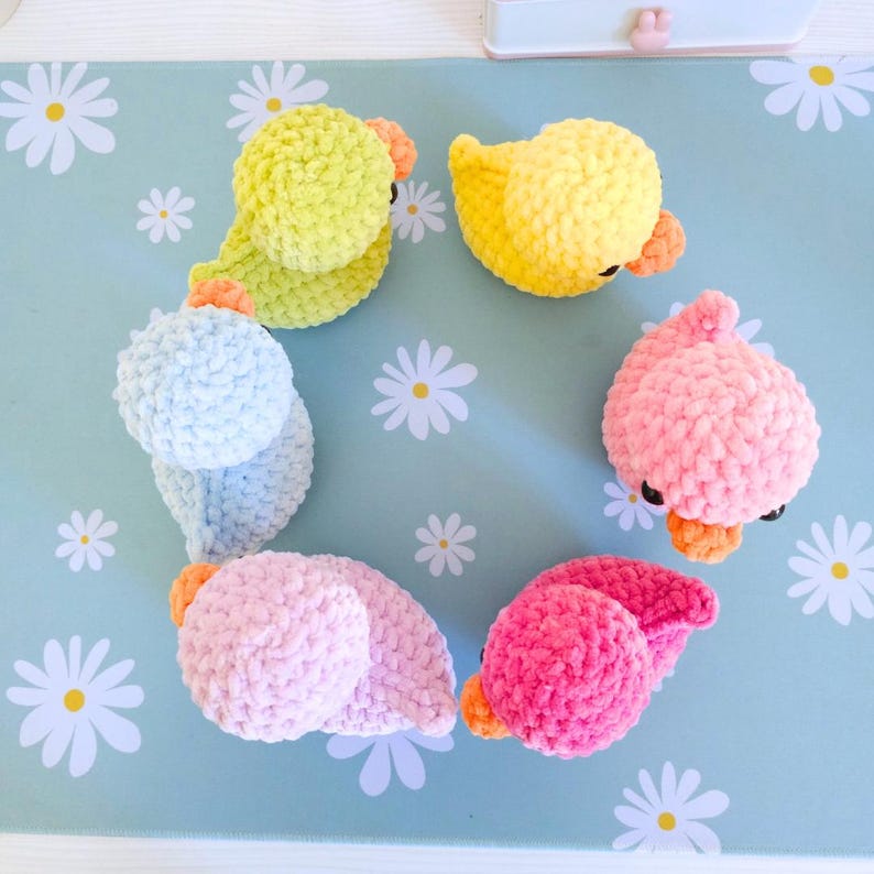 Puede incluir: Siete patitos de ganchillo en amarillo, verde, rosa y azul. Cada patito tiene un pico naranja y ojos negros. Est&aacute;n dispuestos sobre una superficie azul claro con patrones de margaritas blancas.