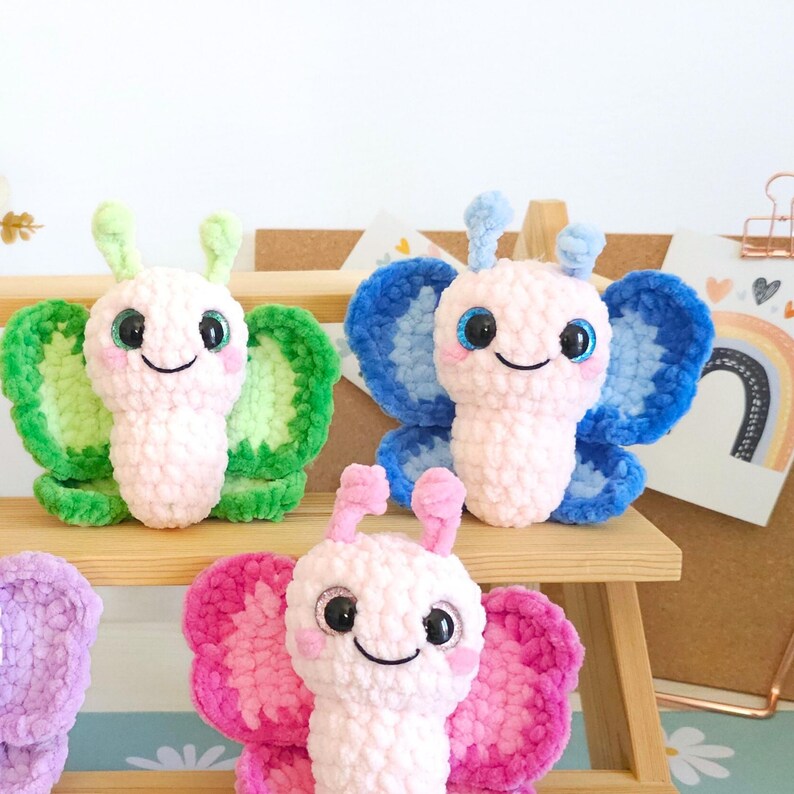 Puede incluir: Juguetes de mariposa de ganchillo en rosa, verde y azul. Cada mariposa de peluche tiene ojos grandes y expresivos y una cara sonriente. Las alas son de varios colores y los juguetes se exhiben en una estanter&iacute;a de madera.