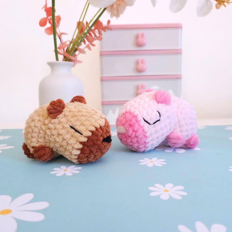 Pu&ograve; includere: Due capibara all'uncinetto fatti a mano. Uno &egrave; beige e marrone, l'altro rosa e bianco. Sono appoggiati su una superficie azzurra con motivi floreali bianchi. Un vaso bianco con fiori rosa e una scatola portaoggetti bianca e rosa sullo sfondo.