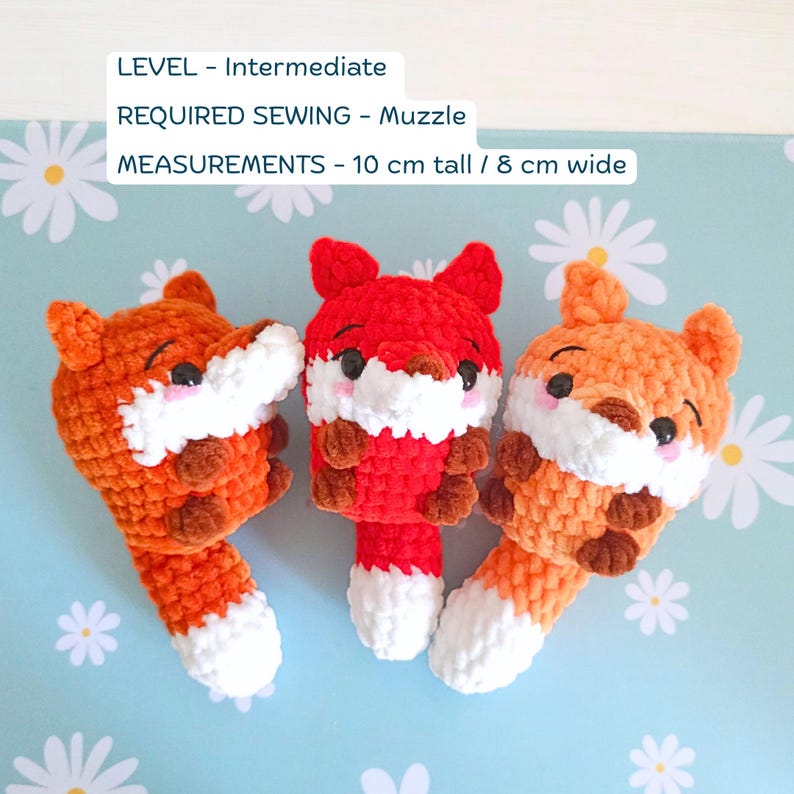 May include: Three crocheted fox toys in shades of orange and red, with white muzzles and tails. The toys are approximately 4 inches tall and 3 inches wide. The text in the image reads: LEVEL - Intermediate, REQUIRED SEWING - Muzzle, MEASUREMENTS - 10 cm tall / 8 cm wide.