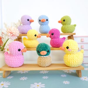 Puede incluir: Una colecci&oacute;n de patitos de ganchillo hechos a mano en varios colores, incluyendo rosa, amarillo, verde, azul y morado. Cada patito tiene un peque&ntilde;o pico naranja y ojos negros. Se exhiben en una estanter&iacute;a de madera.