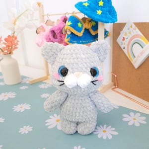 May include: A handmade gray crochet cat doll with blue eyes, pink cheeks, and a white muzzle. The cat is standing in front of a small wooden rack with miniature outfits and a blue wizard hat with yellow stars. The background has a floral pattern.