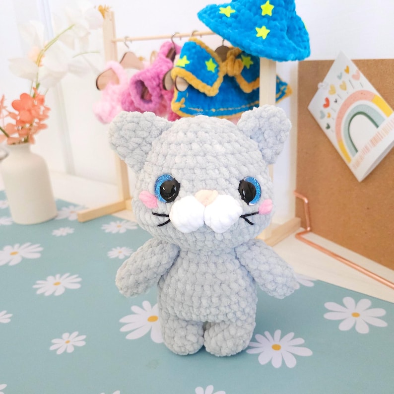 May include: A handmade gray crochet cat doll with blue eyes, pink cheeks, and a white muzzle. The cat is standing in front of a small wooden rack with miniature outfits and a blue wizard hat with yellow stars. The background has a floral pattern.
