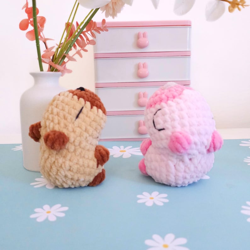 Pu&ograve; includere: Due creature fatte a mano all'uncinetto, una beige e marrone, l'altra rosa e bianca. Sono su una superficie azzurra con motivi floreali bianchi. Un vaso bianco con fiori rosa e una scatola portaoggetti bianca e rosa sono sullo sfondo.