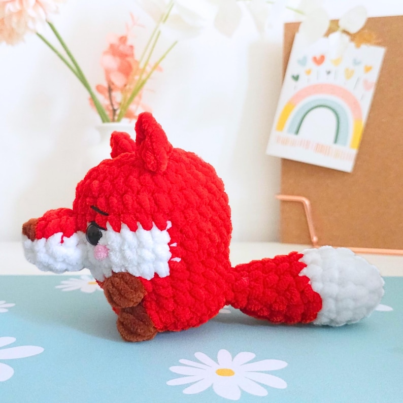 May include: A handmade, red and white crocheted fox toy with a fluffy texture. The fox has a white chest and tail tip, brown accents, and black eyes. The fox is lying on a light blue surface with a daisy pattern.
