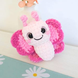 Puede incluir: Un juguete de peluche con forma de mariposa rosa. La mariposa tiene ojos grandes y brillantes, una boca sonriente y peque&ntilde;as antenas. Las alas son de un rosa m&aacute;s oscuro con detalles en rosa m&aacute;s claro. El juguete es suave.