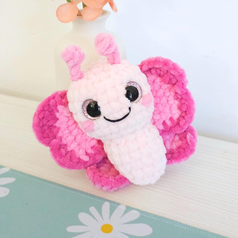 Puede incluir: Un juguete de peluche con forma de mariposa rosa. La mariposa tiene ojos grandes y brillantes, una boca sonriente y peque&ntilde;as antenas. Las alas son de un rosa m&aacute;s oscuro con detalles en rosa m&aacute;s claro. El juguete es suave.