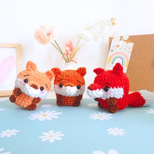 May include: Three crocheted fox toys in shades of orange and red, with white and brown accents. The foxes have small ears, black eyes, and white muzzles. One fox is lying down, while the others are upright. The toys are on a blue surface with white flower patterns.