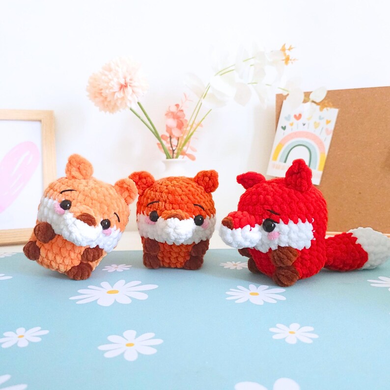 May include: Three crocheted fox toys in shades of orange and red, with white and brown accents. The foxes have small ears, black eyes, and white muzzles. One fox is lying down, while the others are upright. The toys are on a blue surface with white flower patterns.