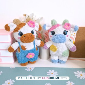 May include: Two handmade crochet cow plushies. One cow is white and brown with blue overalls and a pink flower. The other is white with pastel accents and a pink flower. The text "PATTERN BY HUGURUMITOYS" is at the bottom.